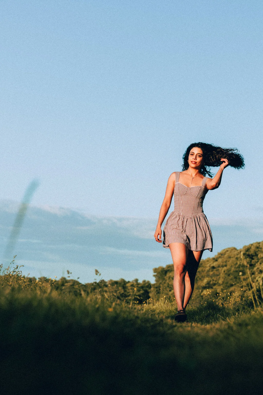 A woman with curly hair in a gingham dress walking through a grassy field under a clear blue sky.