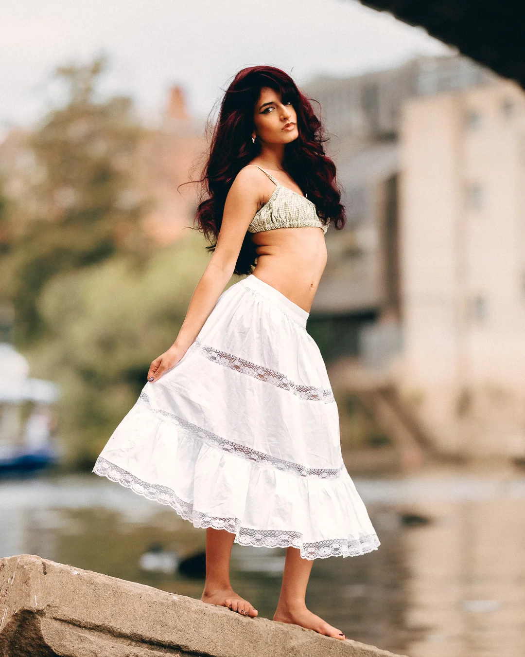 Woman with long red hair in a gold crochet top and white lace skirt posing on a rock by the water.