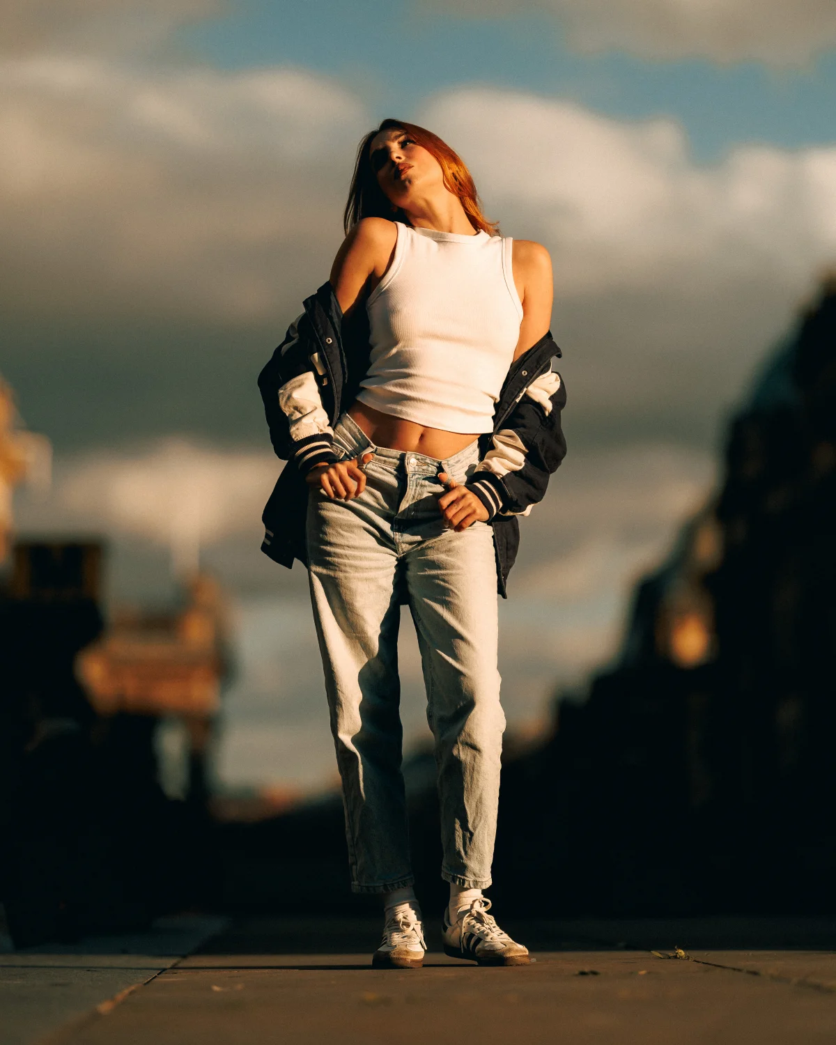 A young woman with red hair posing in a white crop top, light-wash jeans, and a varsity jacket under a cloudy sky.