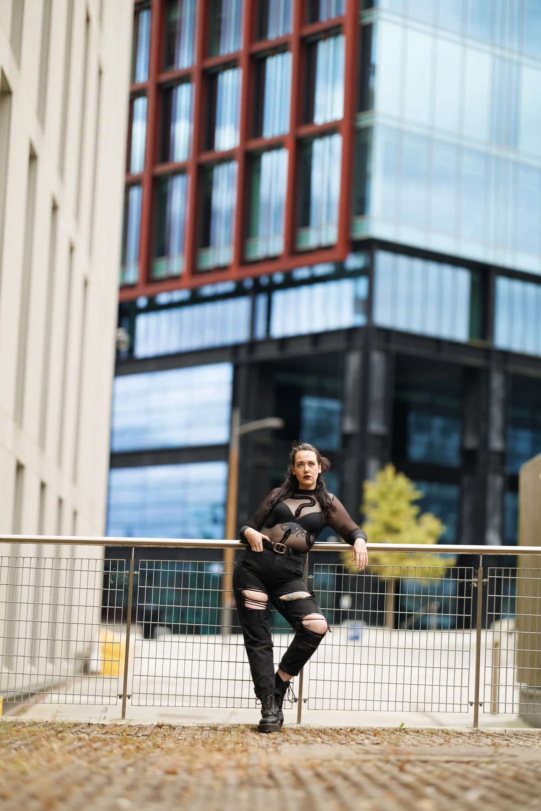 Woman in a sheer black top and ripped black jeans posing by a railing in an urban city setting.
