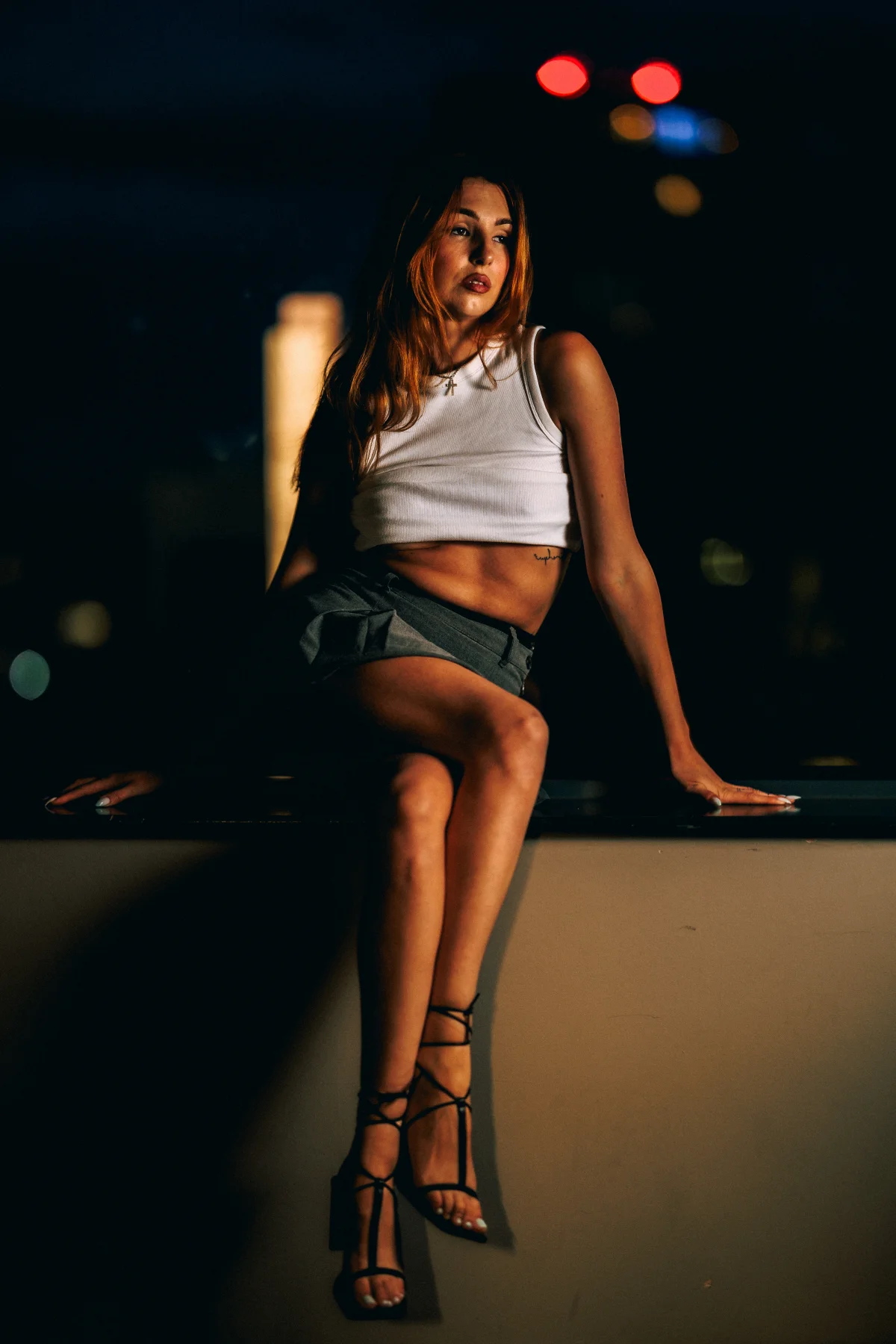 A woman in a white crop top and grey shorts posing on a ledge at night with blurred city lights in the background.