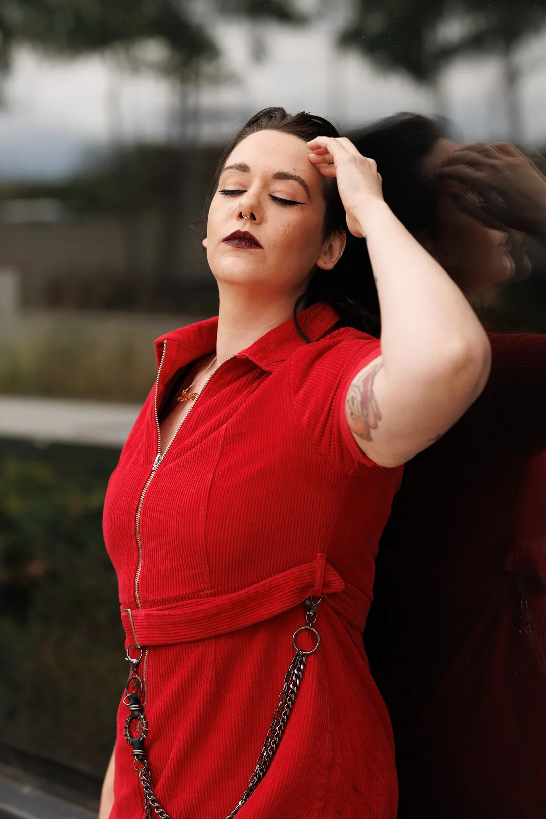 A woman in a red corduroy dress with chains, eyes closed, leaning against a reflective surface.