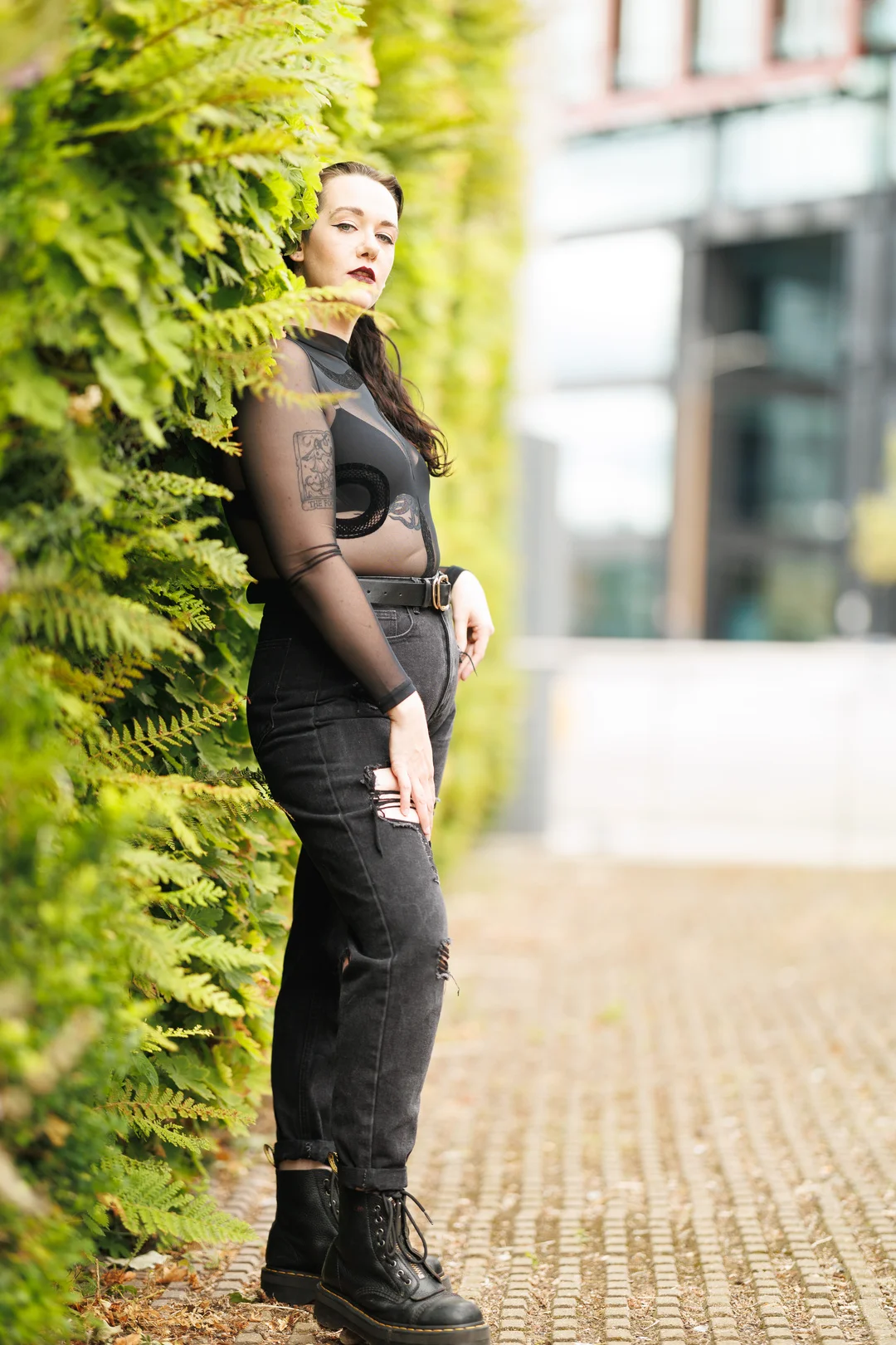 Woman in a black mesh top and ripped black jeans posing against a lush green hedge outdoors.