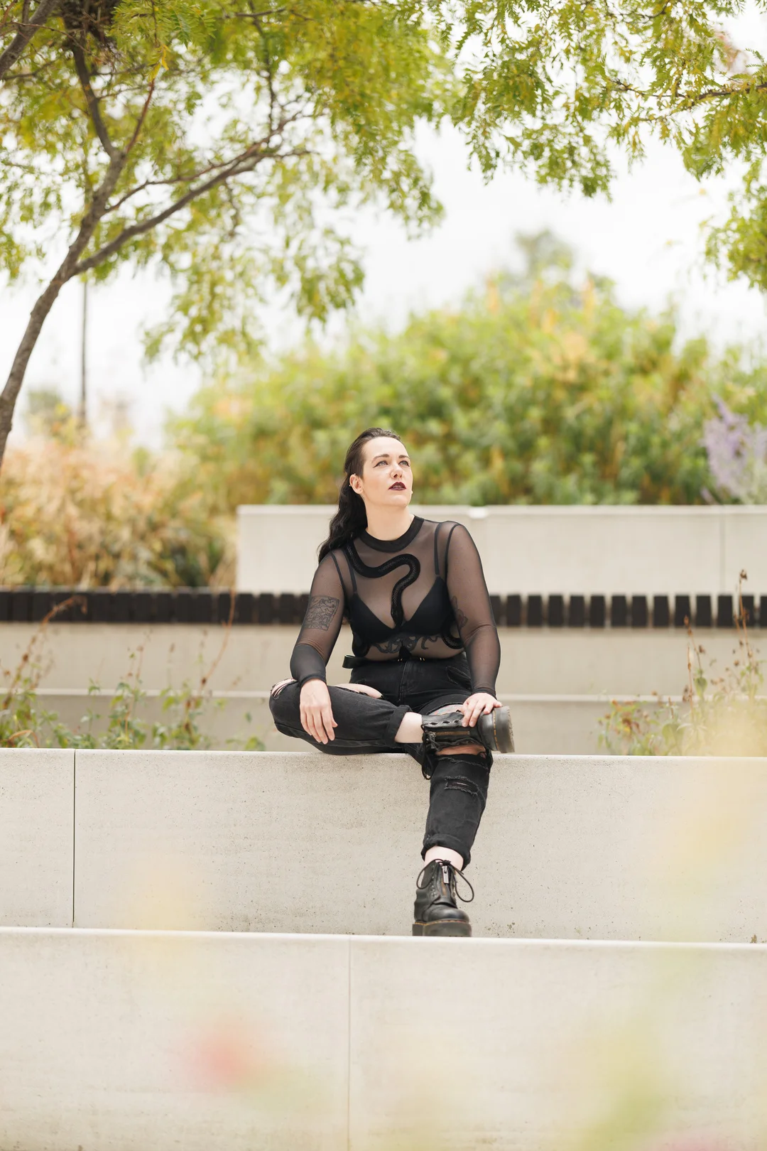 Person with dark makeup and a sheer black top sitting on a concrete ledge in a garden setting.