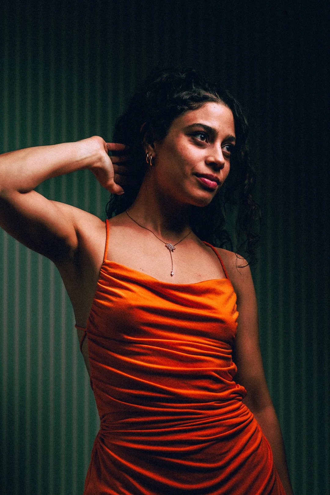A woman with curly dark hair in an orange satin dress posing against a dark green striped background.