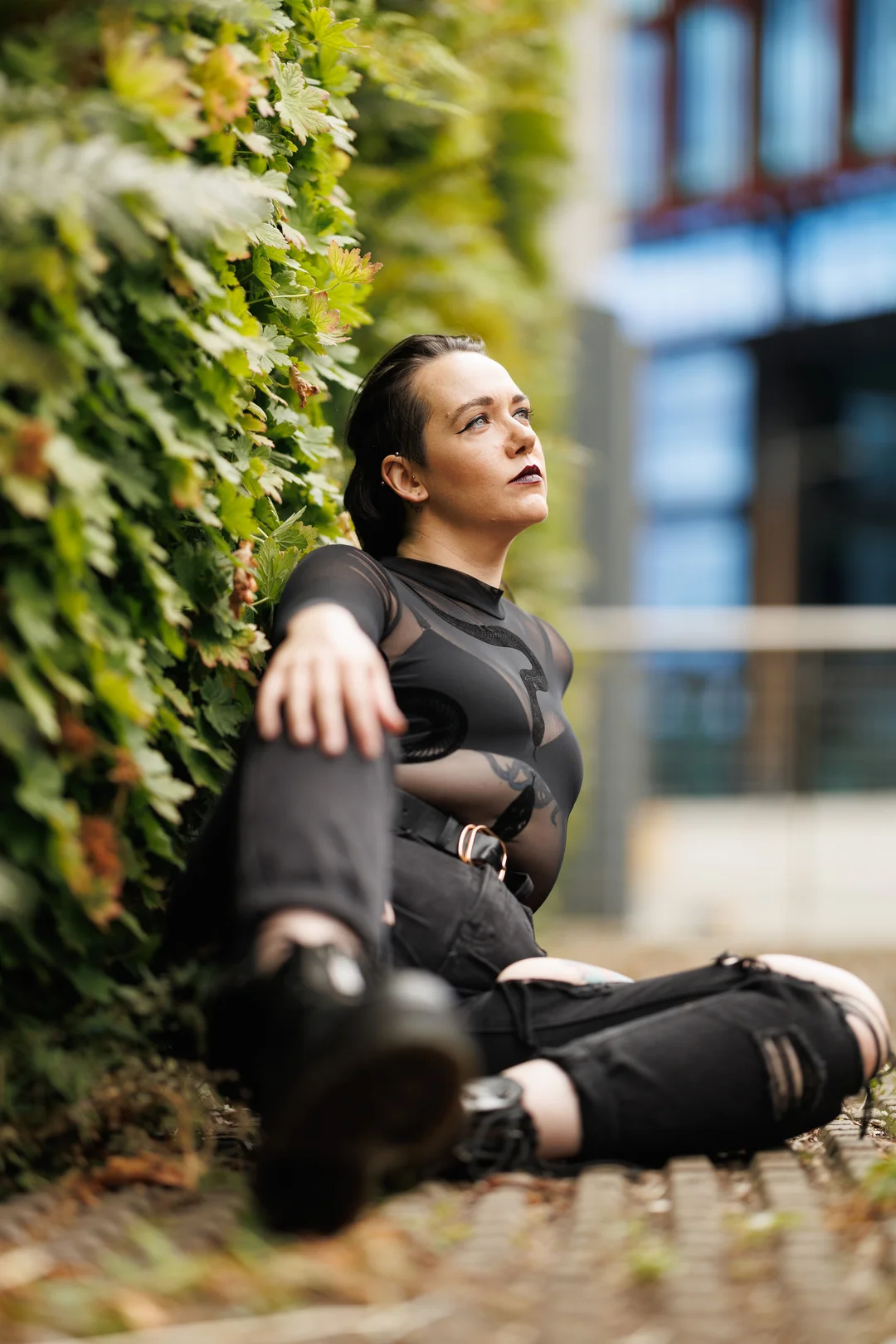 A person in a black sheer top and ripped jeans sitting against a green wall, looking away.