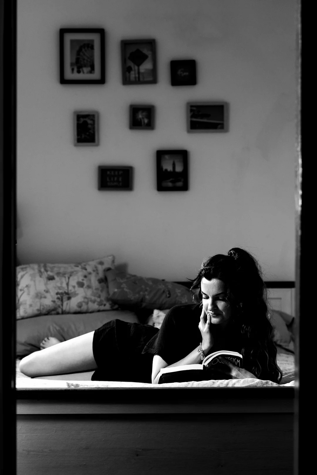 Black and white photo of a woman reading a book while lying on a bed in a moody room.