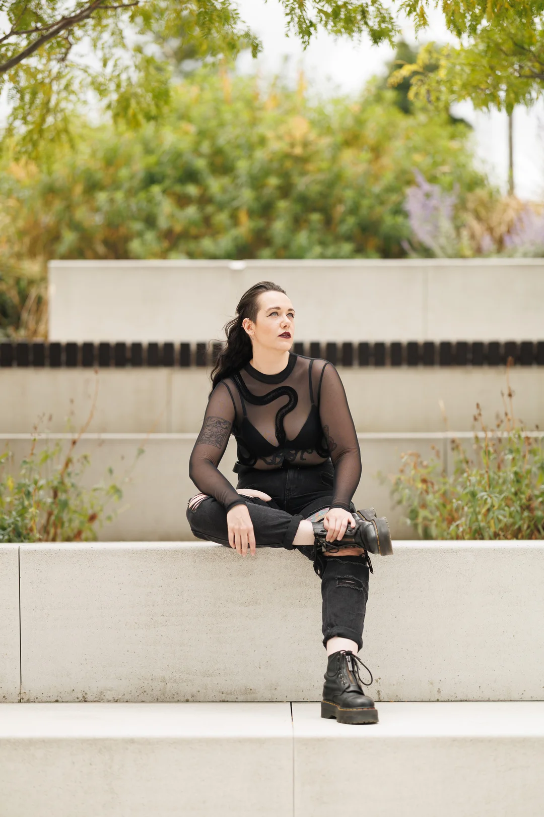 Woman with dark hair and makeup in a black mesh top and ripped jeans sitting on a concrete ledge outdoors.