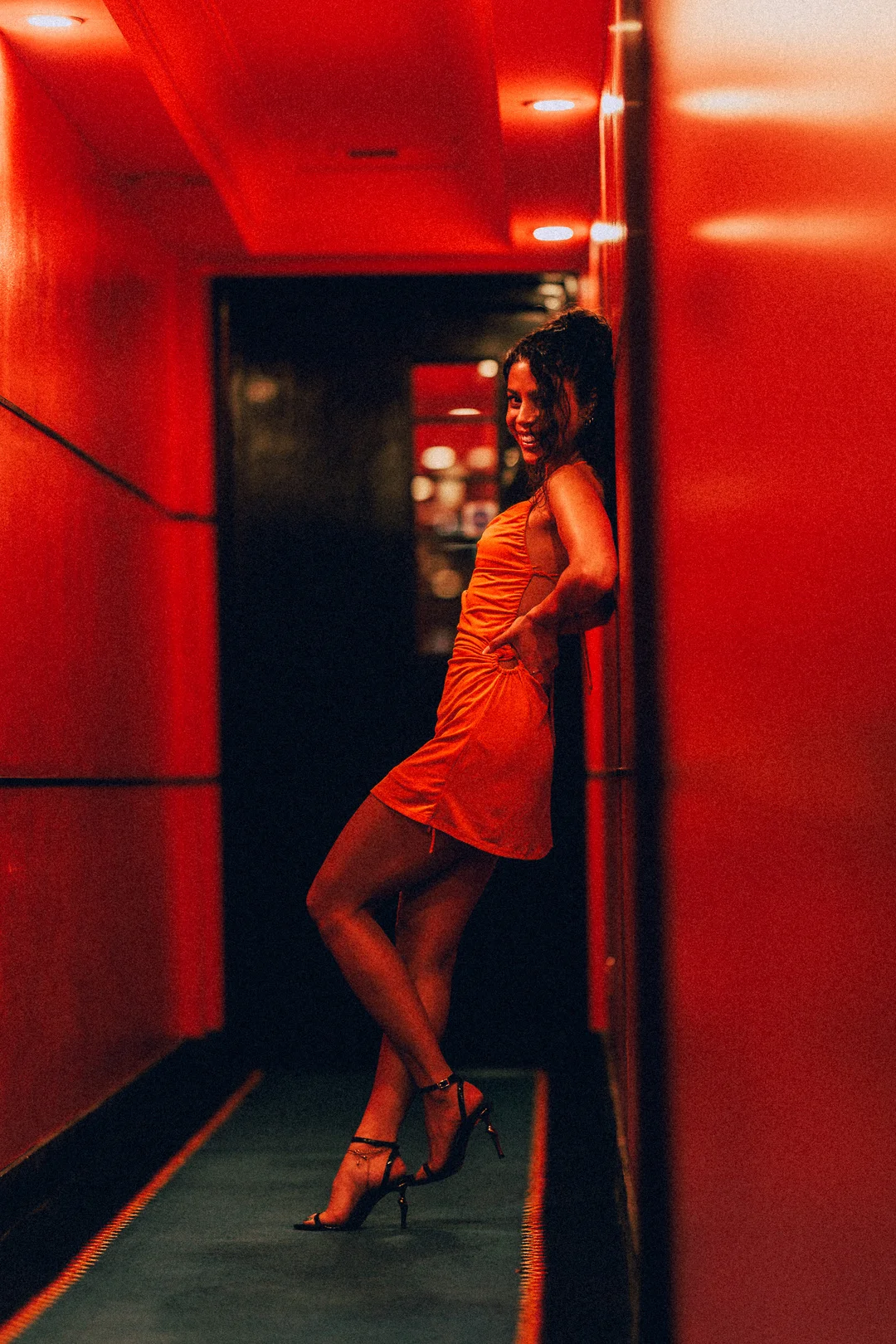 Woman in a short orange dress and high heels posing in a red-lit hallway
