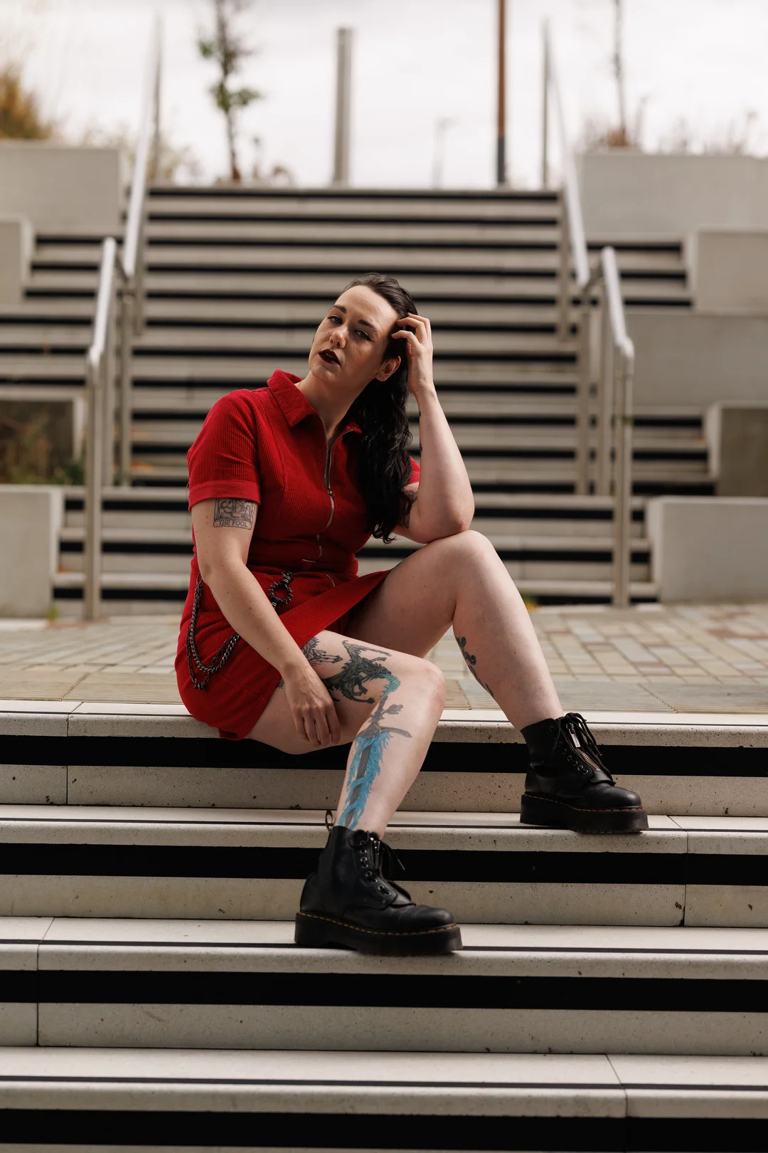 Woman with tattoos in a red dress and black boots posing on outdoor concrete stairs.