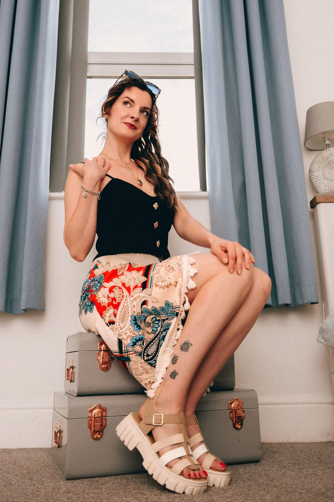 Woman in a black top and colorful paisley skirt posing on grey suitcases by a window.