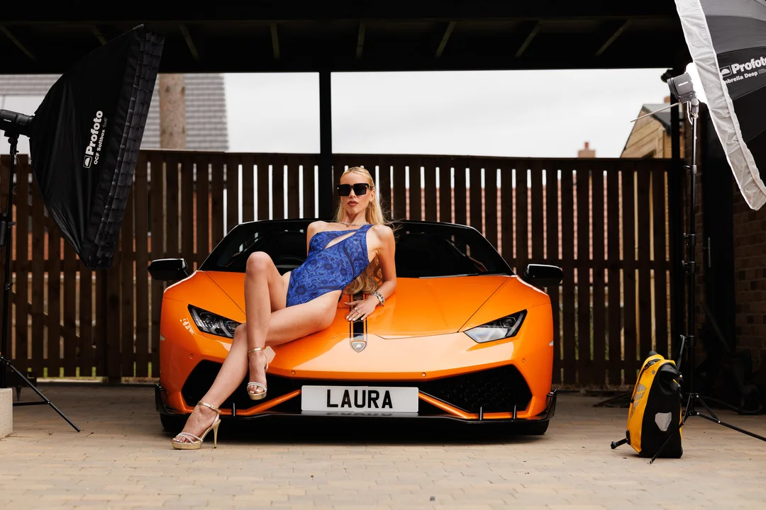 Woman in a blue swimsuit and sunglasses posing on an orange Lamborghini with studio lights.