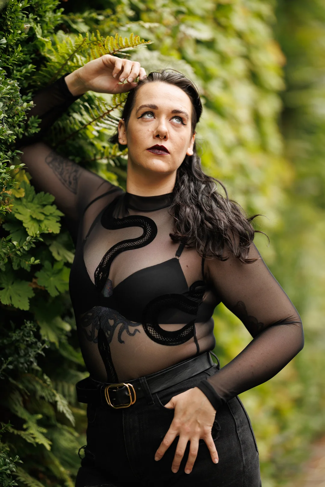 A woman with dark hair and bold makeup posing against a lush green hedge in a sheer black snake-print top.