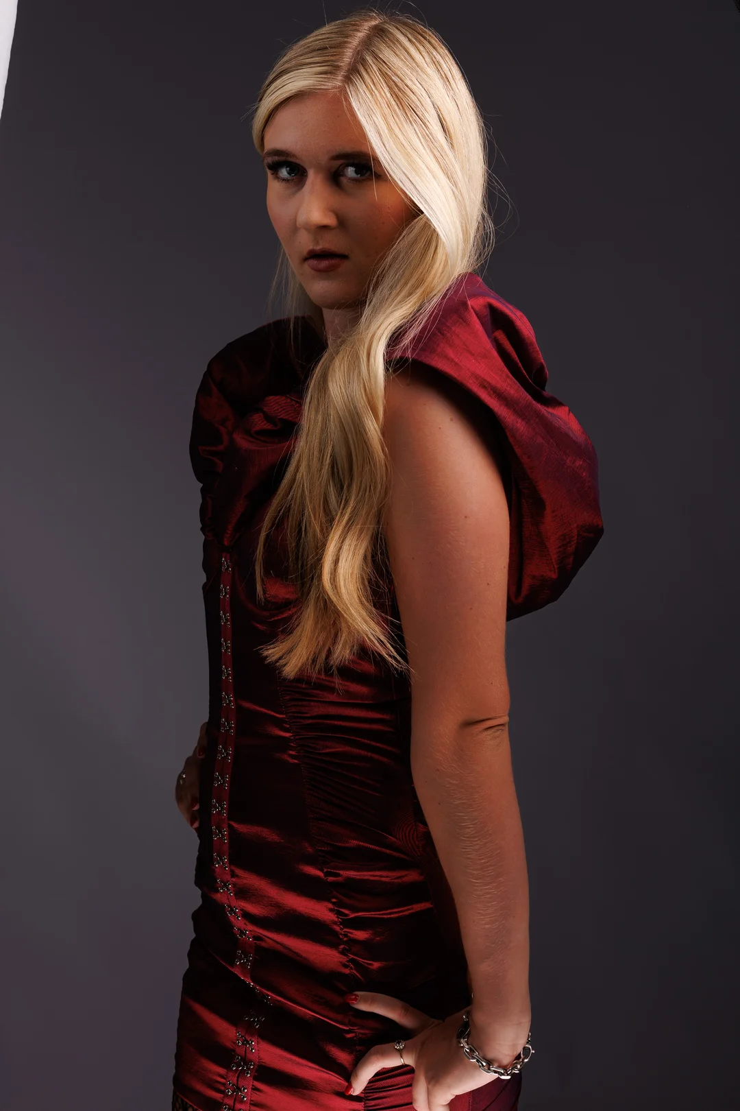 Blonde woman in a shimmering red dress posing against a dark gray studio background.
