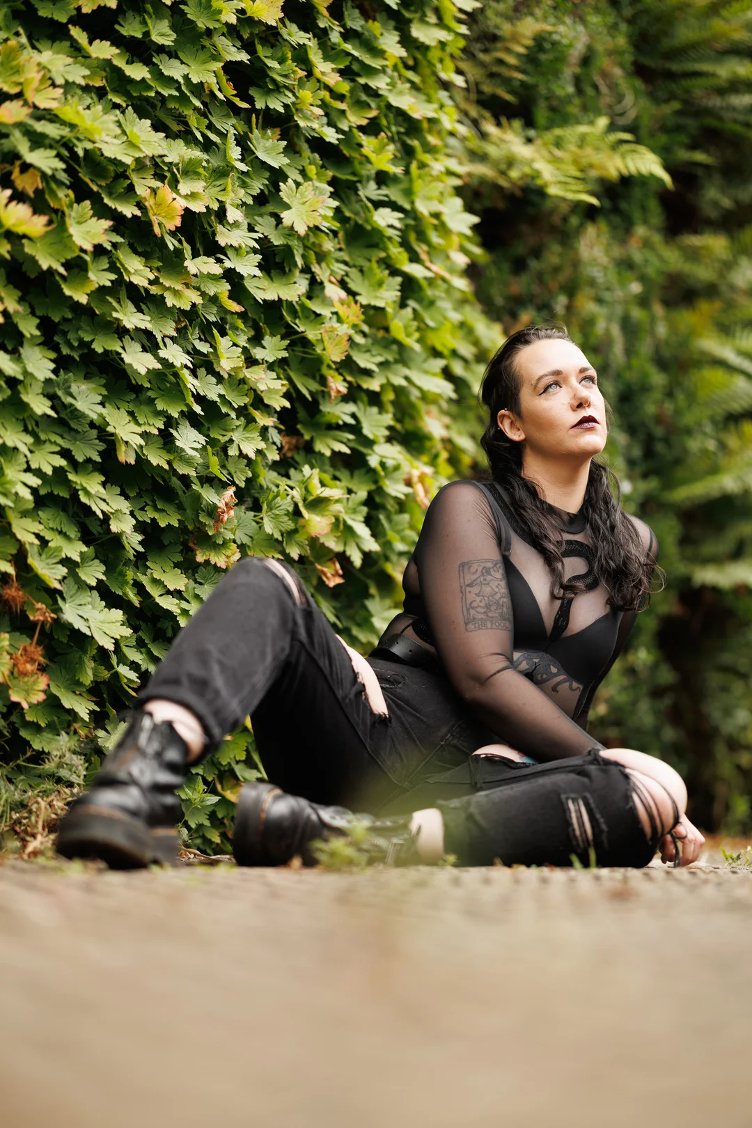 A person with dark hair and makeup, wearing a black mesh top and ripped black jeans, sitting against a green hedge.