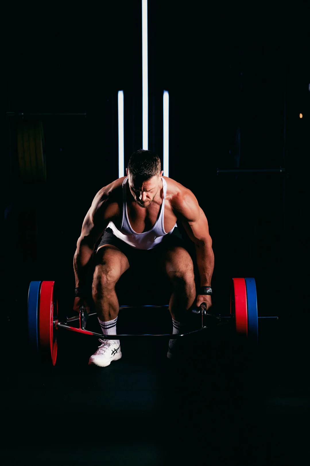Muscular man in a white tank top lifting a trap bar in a dark studio with dramatic vertical lighting.