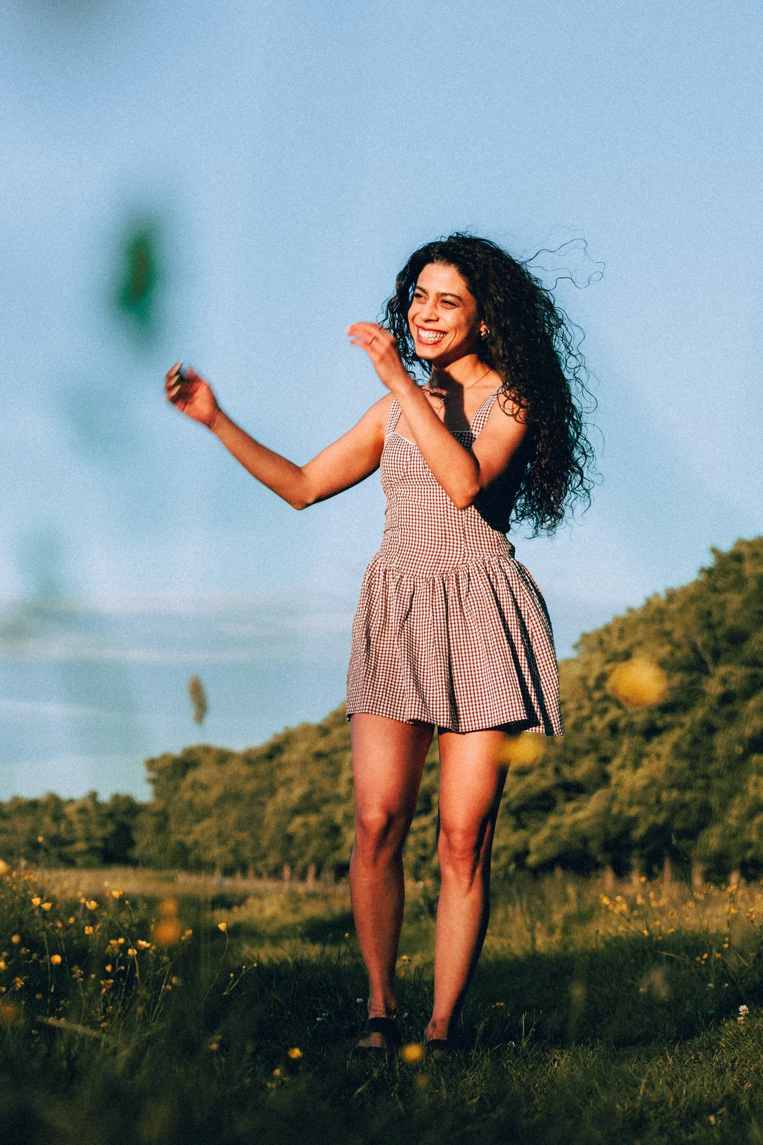 A smiling woman with curly hair in a red and white gingham dress standing in a sunny meadow.