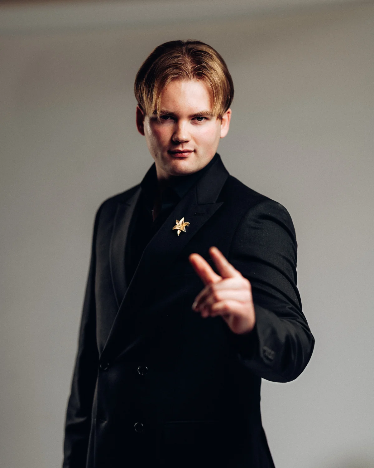 Confident man in a black suit pointing at the camera against a plain grey background.