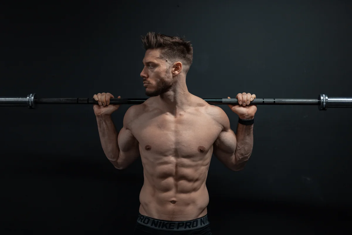 Muscular man posing shirtless with a barbell across his shoulders against a dark studio background.