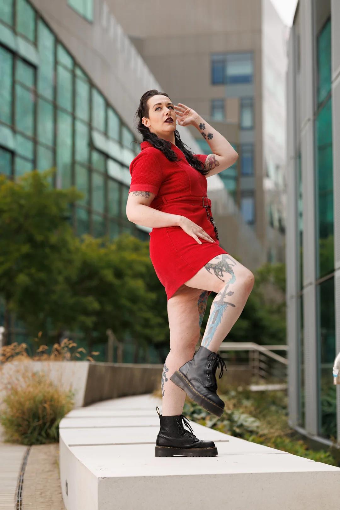 A woman with tattoos in a red dress and black boots posing in an urban outdoor setting.