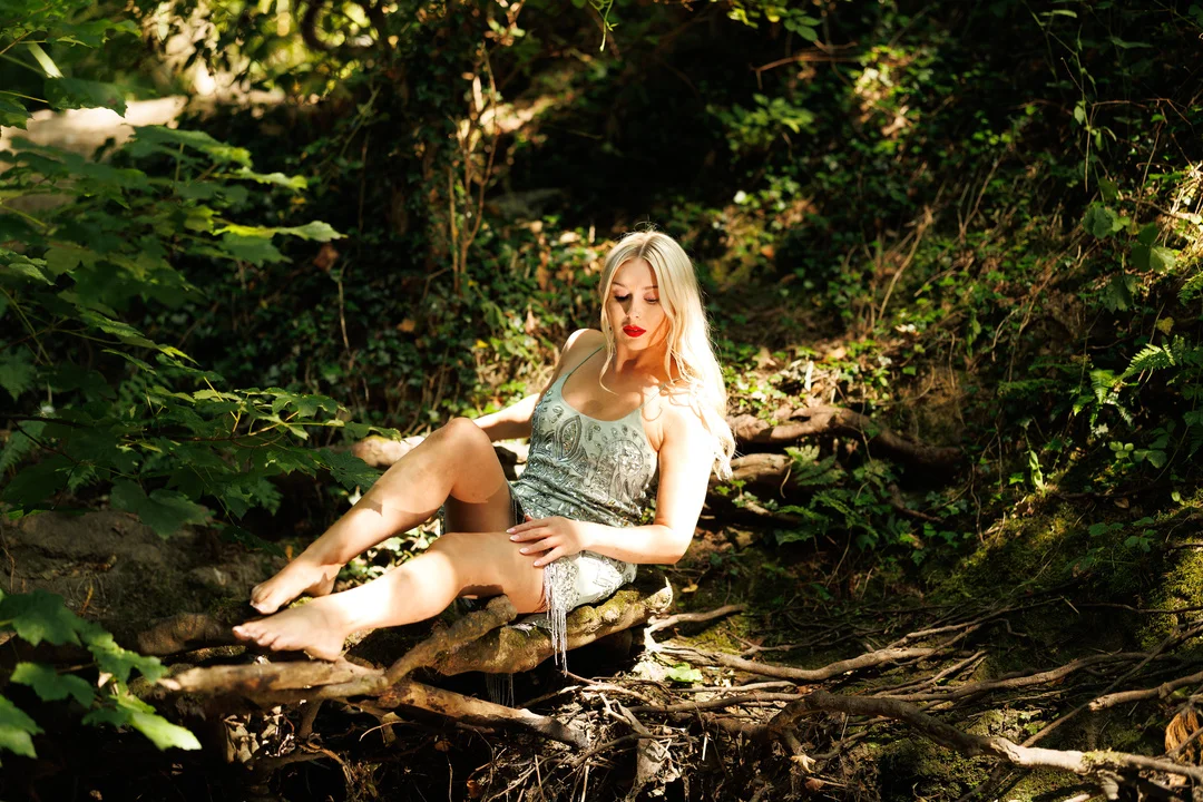 Blonde woman in a silver sequined dress posing on a log in a lush green forest.
