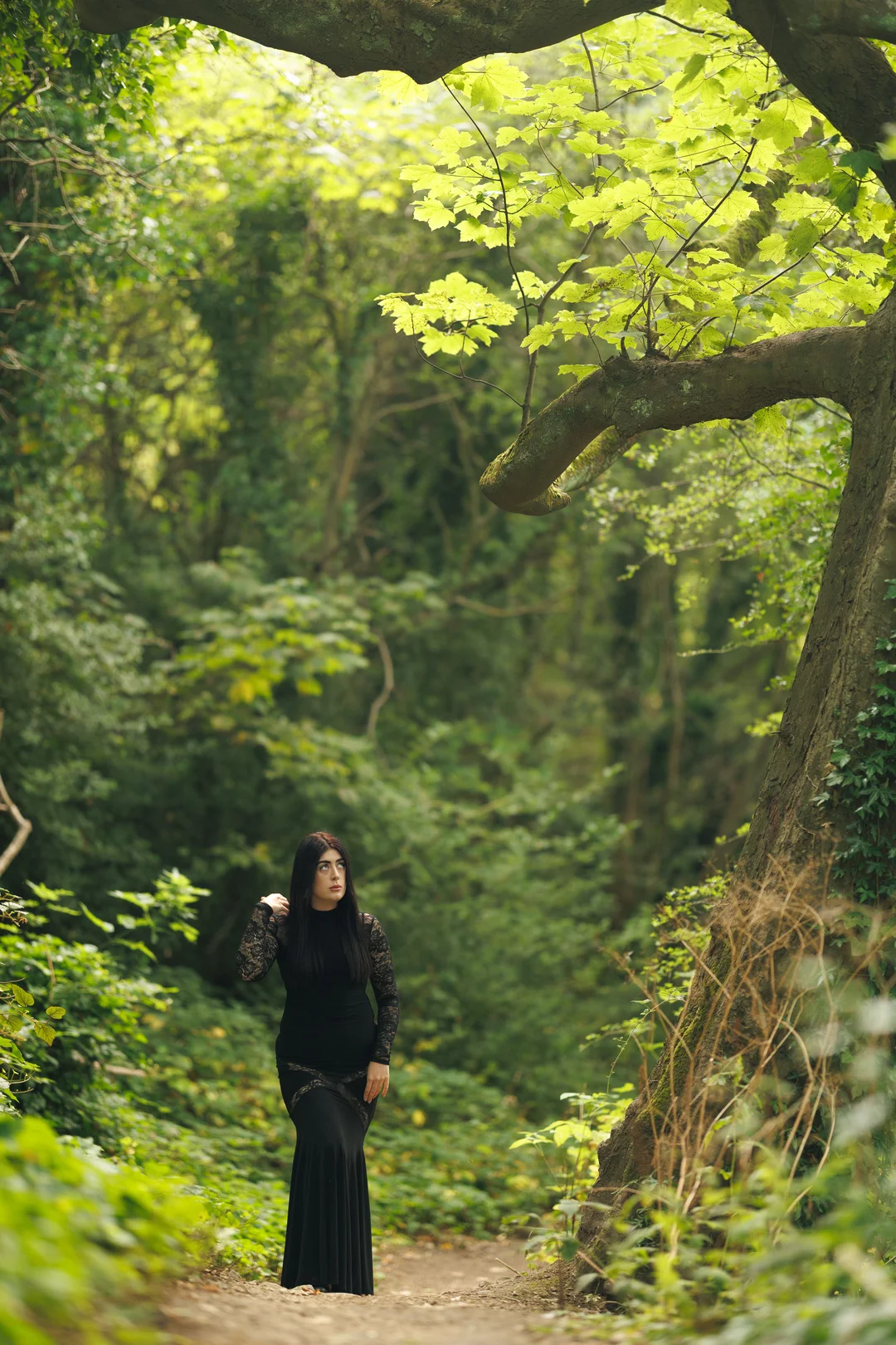 Woman in a long black lace dress standing on a forest path, looking thoughtfully to the side.