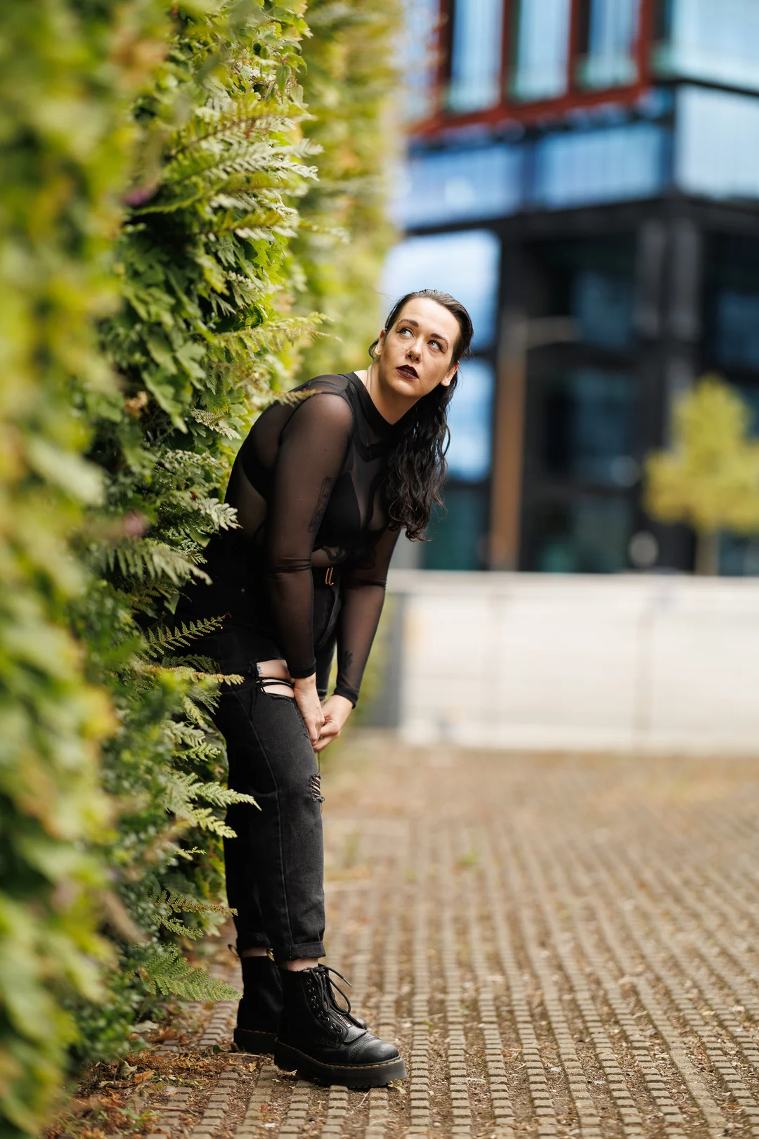 A woman with dark hair and bold makeup posing against a green hedge in an urban setting, wearing a black mesh top and ripped jeans.