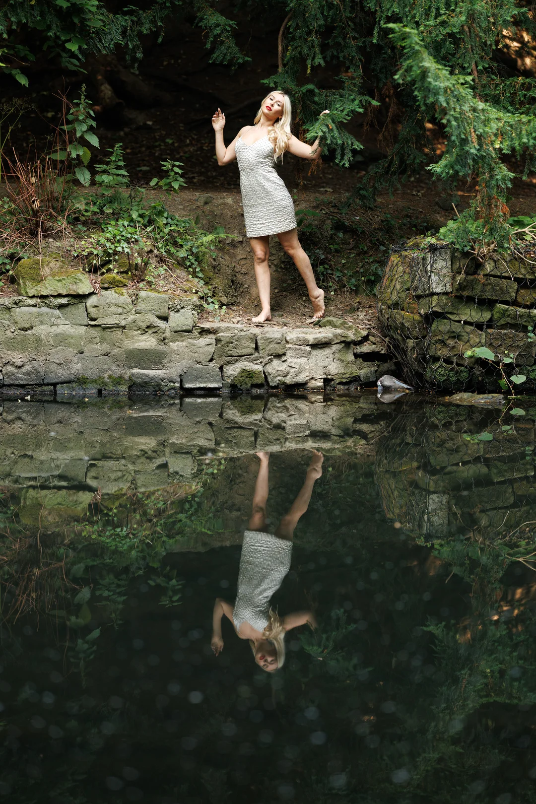 Woman in a silver dress posing on a stone ledge with a clear reflection in a still pond.