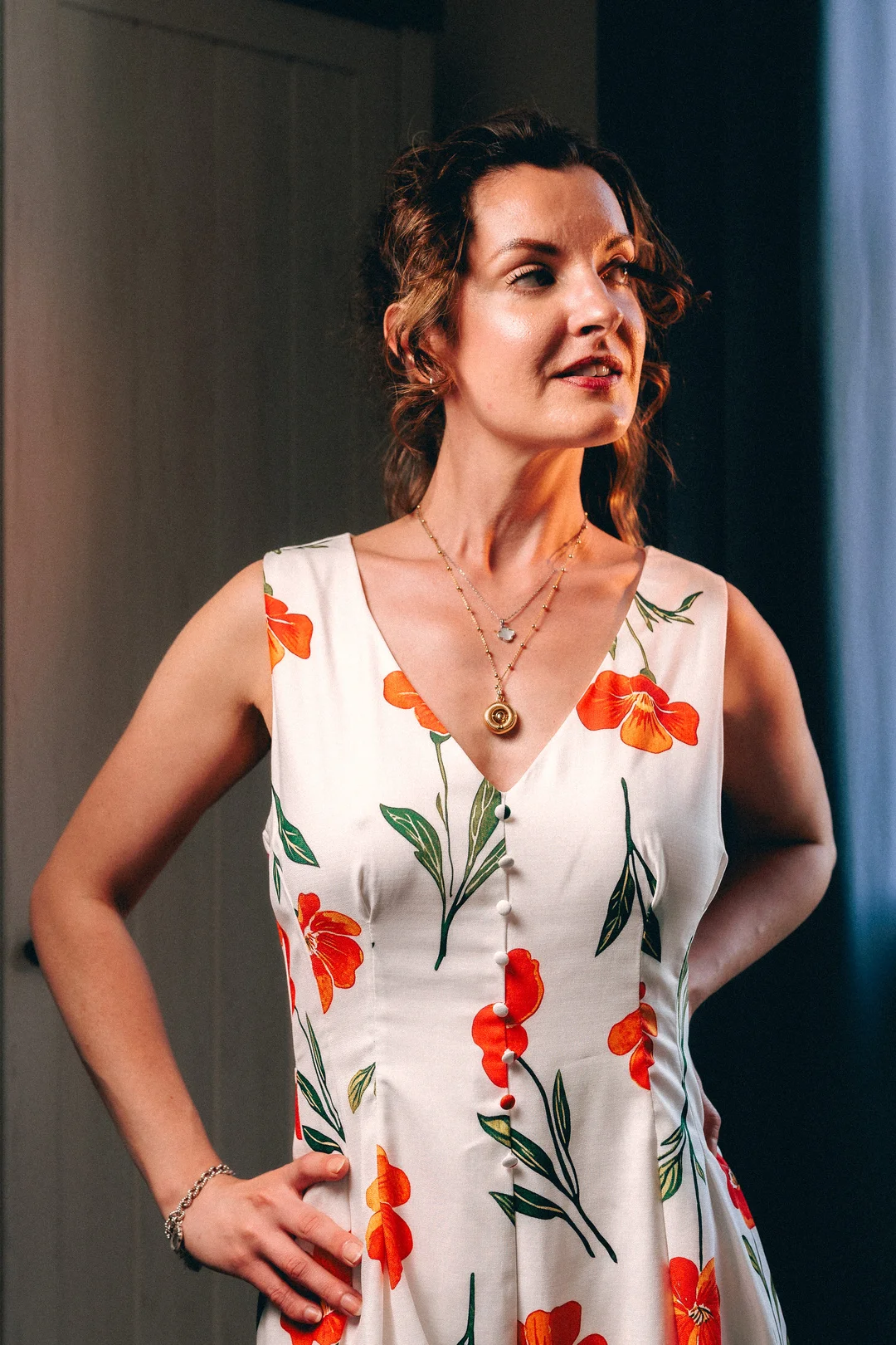 A woman with curly brown hair in a white floral dress, looking thoughtfully to the side in soft natural light.