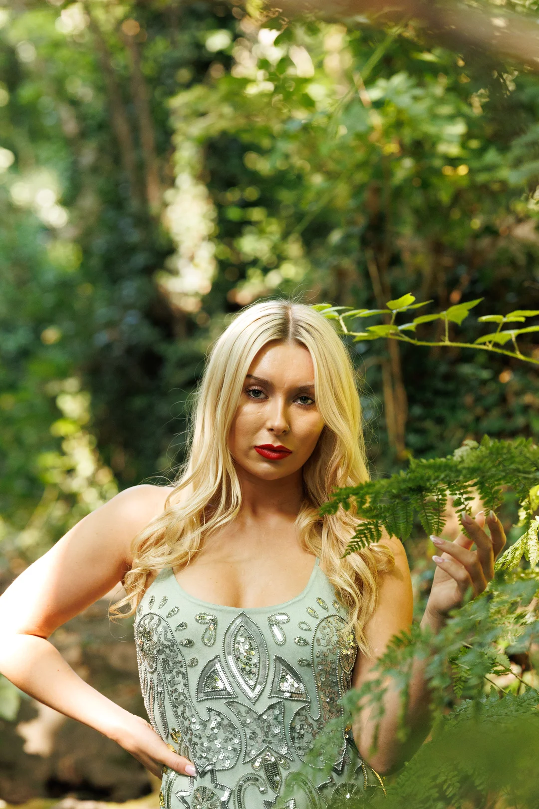 A blonde woman in a sequined light-green dress posing in a lush green forest.