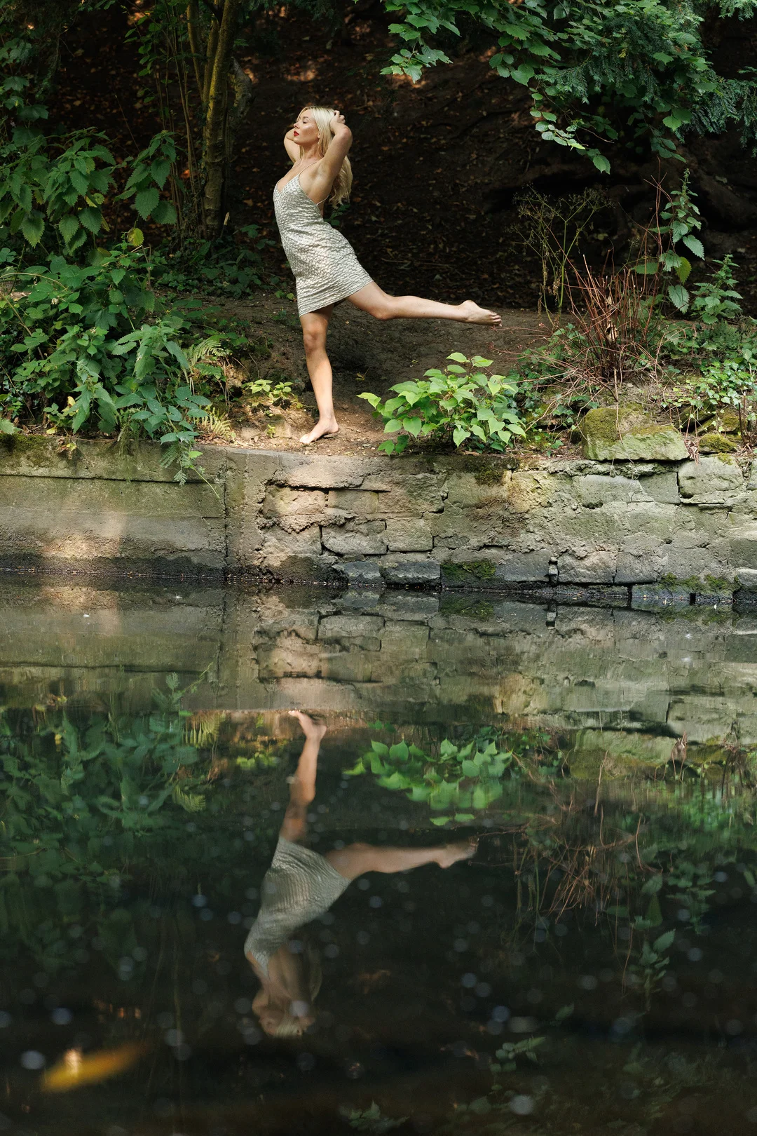 Woman in a short gold dress posing on a stone bank with a reflection in the water below.