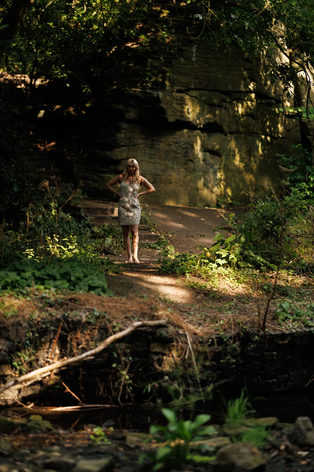 A woman in a short, sparkly dress posing in a wooded area with a rocky cliff background.