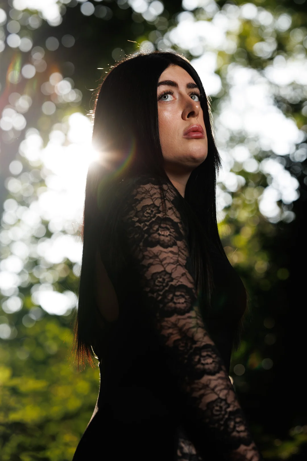 Portrait of a woman with long dark hair in a black lace dress, looking up with sunlight filtering through trees.