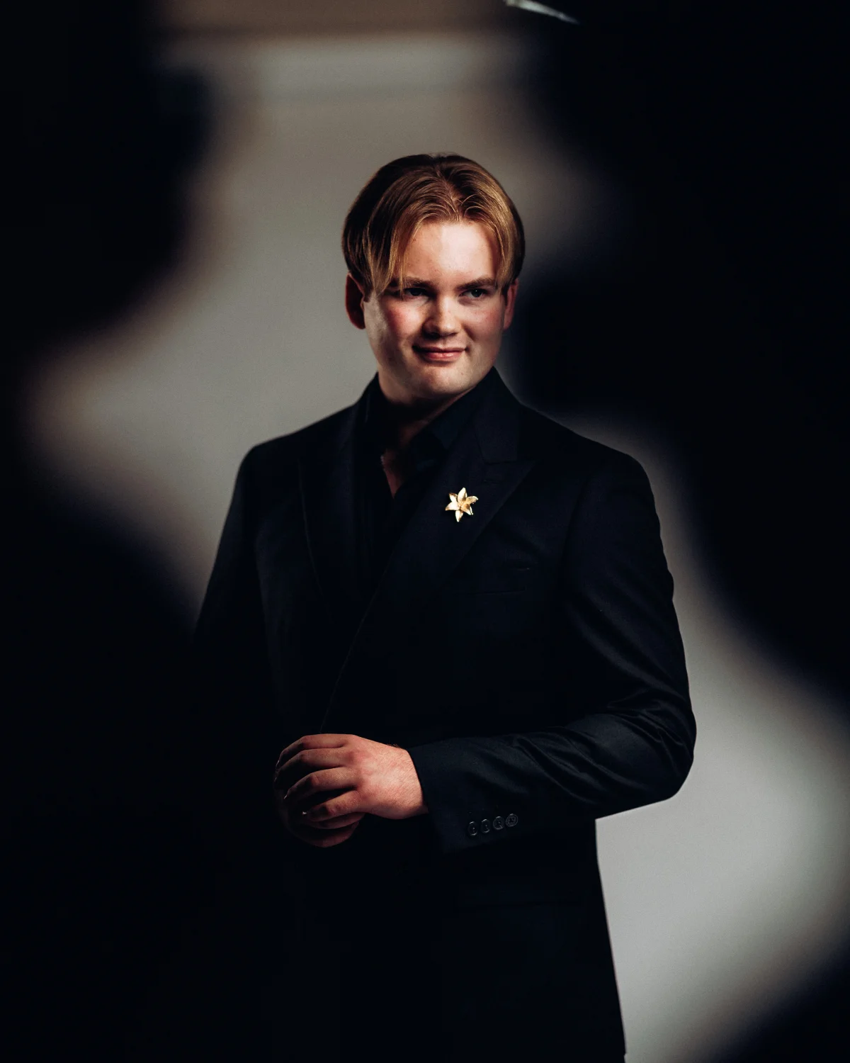 A young man with reddish-blonde hair in a black suit and gold lapel pin, posing against a dark, moody background.