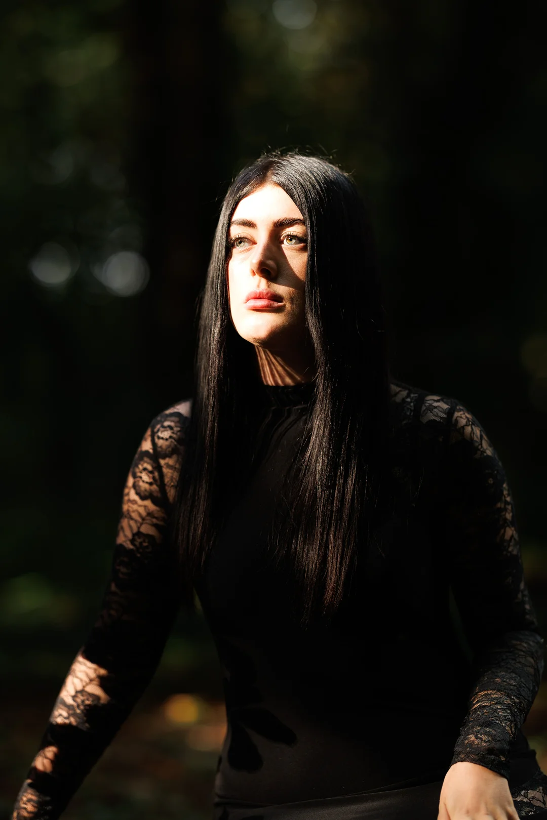 Moody portrait of a woman with long black hair in a black lace top, lit by natural light in a dark forest.