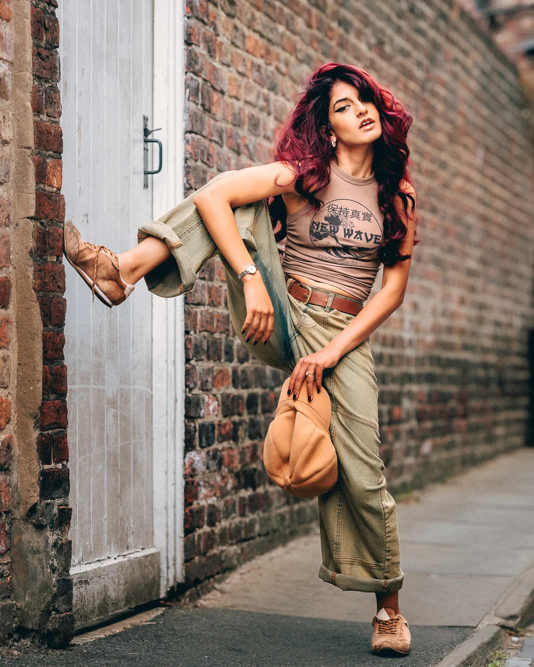 Woman with red hair posing in an urban alley wearing a tan crop top and green pants.
