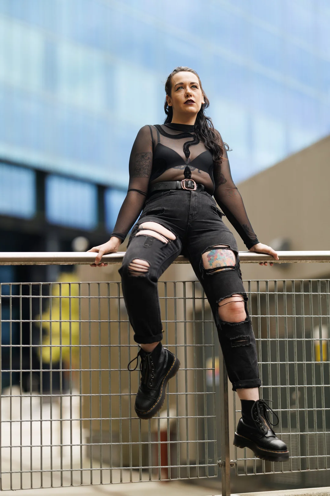 A woman in an edgy black sheer top and ripped jeans posing on a metal railing in an urban setting.