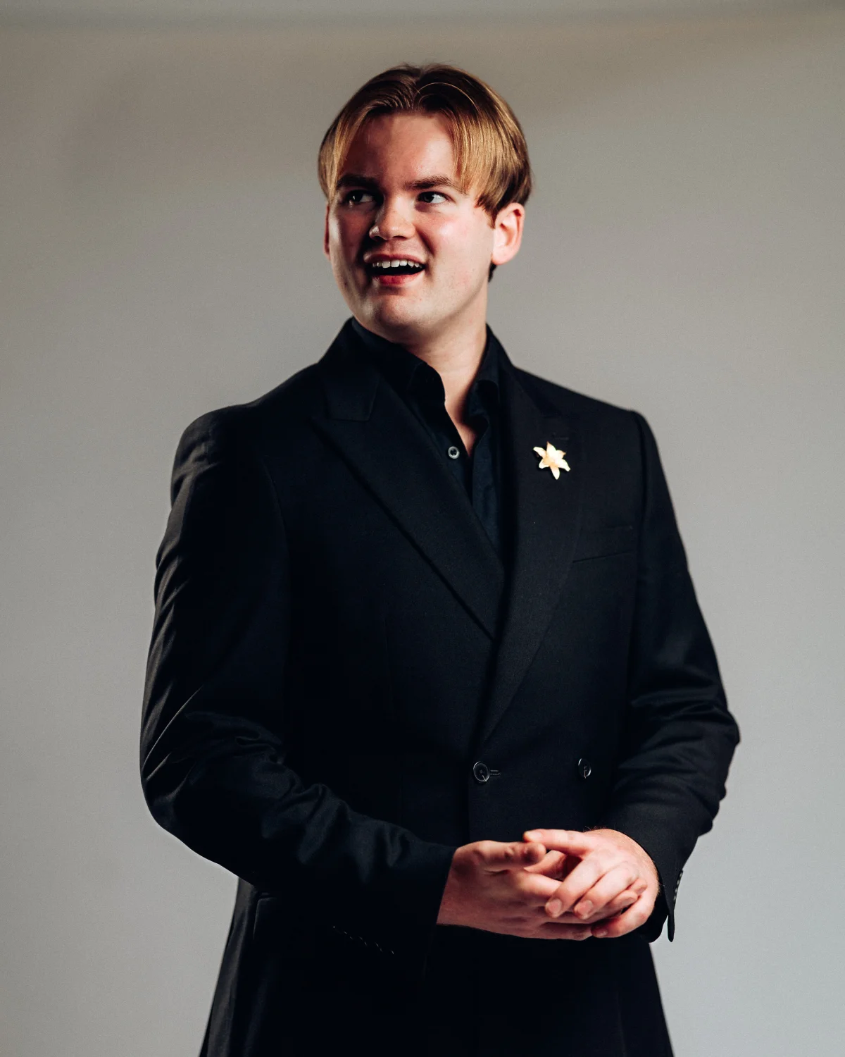 A professional studio portrait of a smiling young man in a black suit against a neutral grey background.