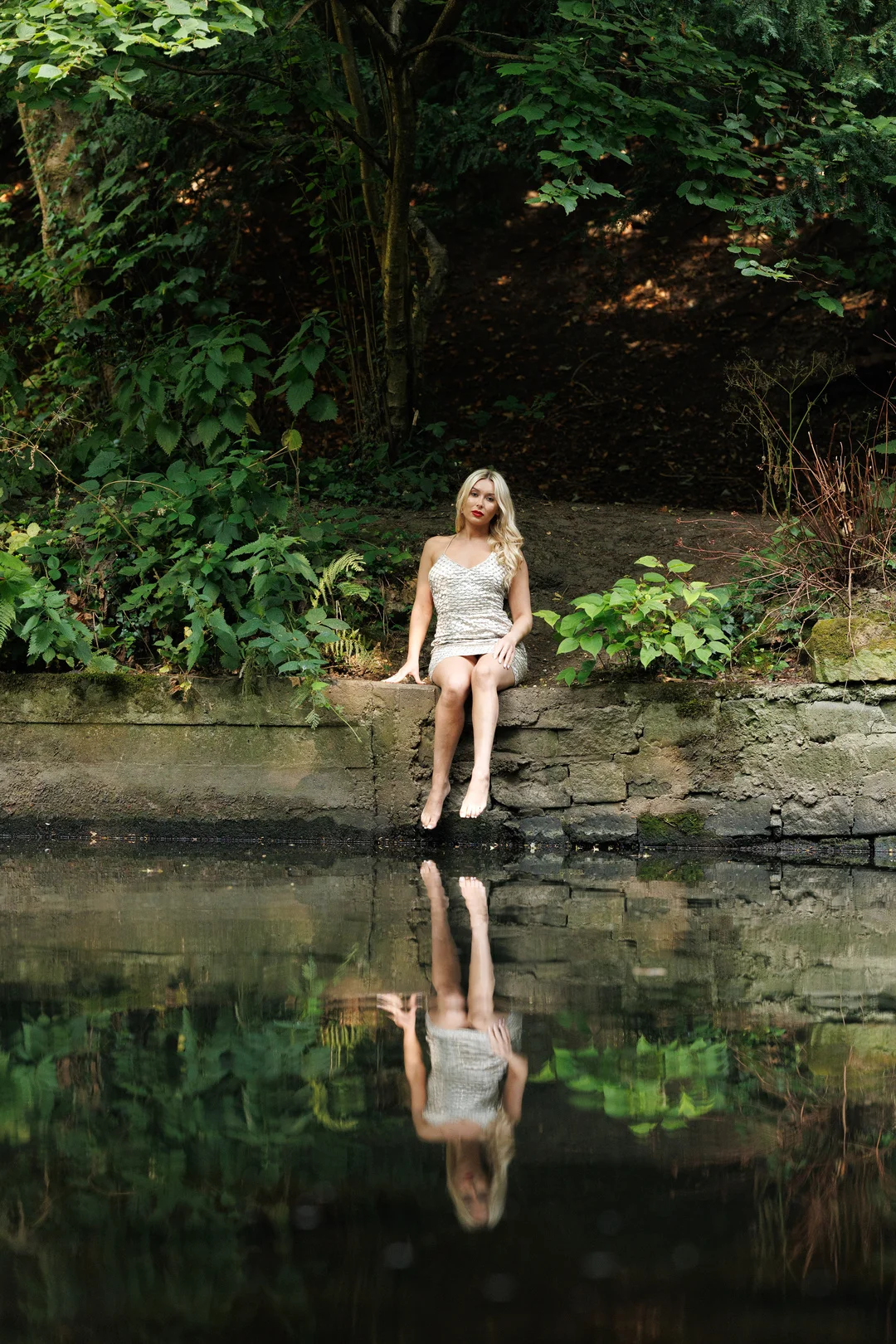 Blonde woman in a short sparkly dress sitting on a stone ledge by a pond with a clear reflection in the water.