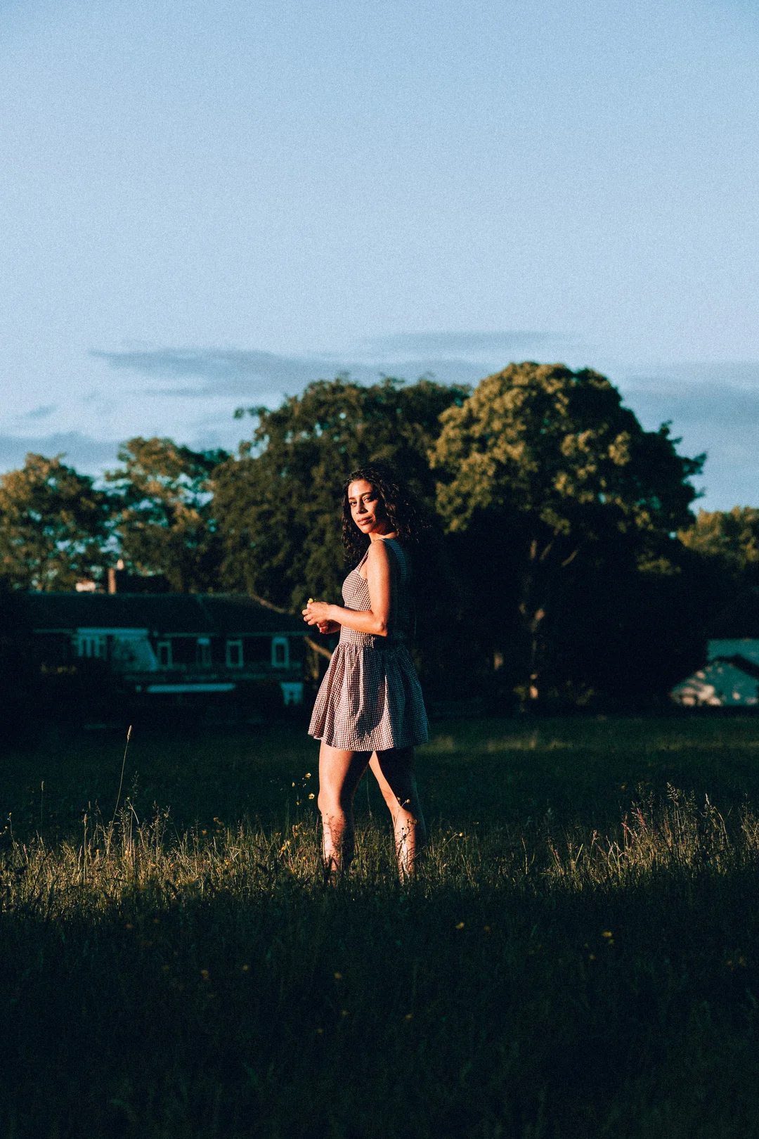 A woman in a red and white checkered dress standing in a grassy field during golden hour.