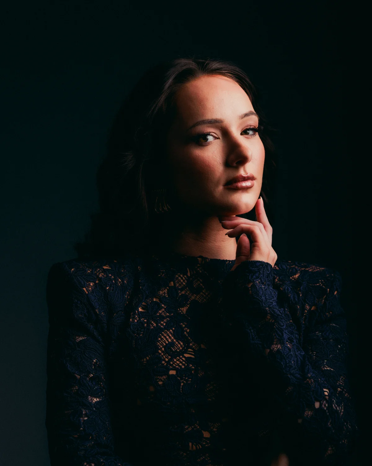 Moody studio portrait of a woman in a dark lace dress posing against a black background.