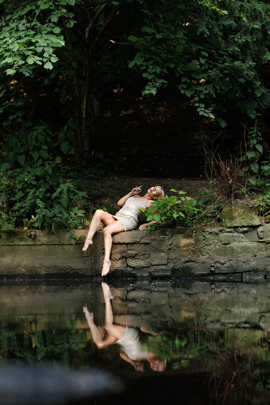 Woman in a shimmering silver dress posing on a stone wall by a pond with a clear reflection.