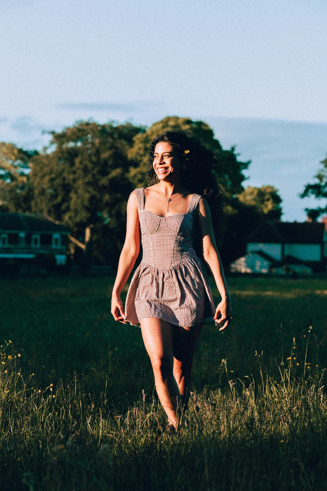 Smiling woman with curly hair in a red gingham dress walking through a sunny grassy field.