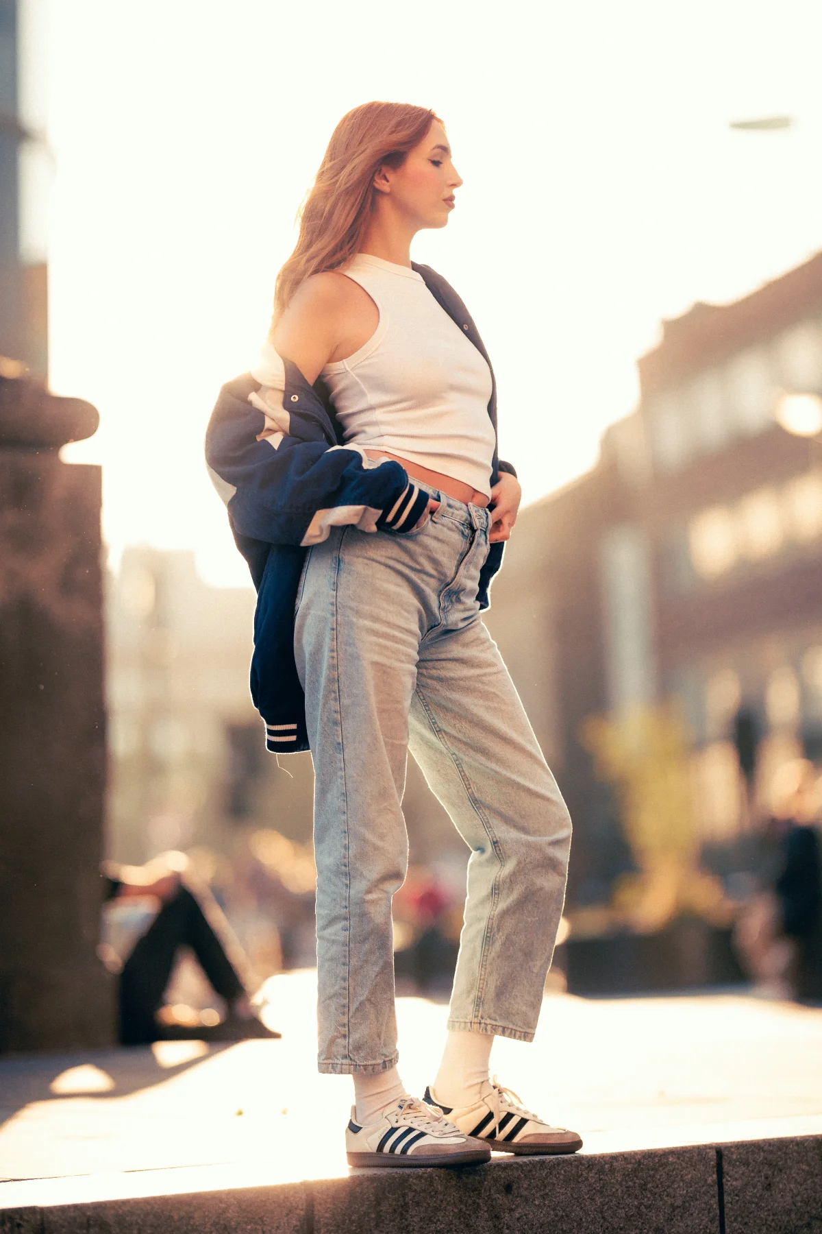 Full-body shot of a red-haired woman in a white tank top, light-wash jeans, and a navy varsity jacket in an urban setting.