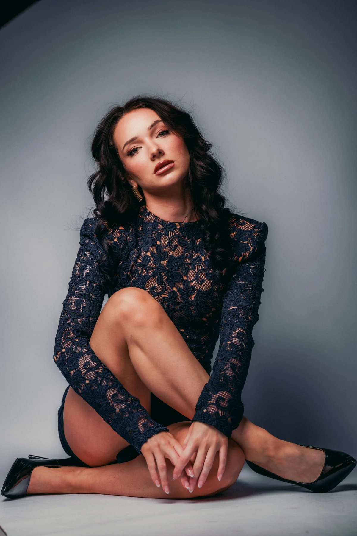 Woman in a navy blue lace dress and black heels posing against a grey studio backdrop.