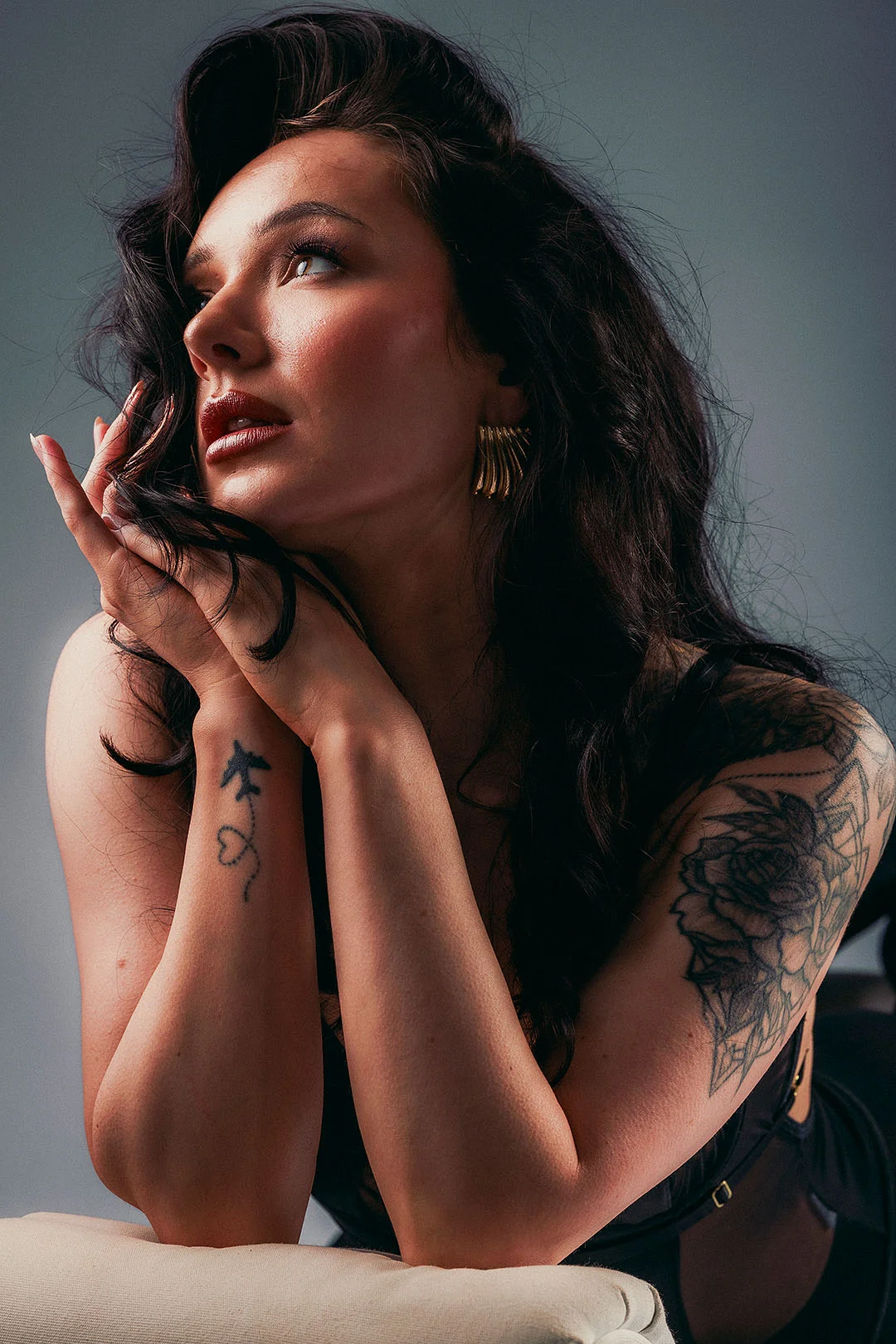 Close-up portrait of a woman with dark wavy hair and tattoos, looking upwards with a moody expression.