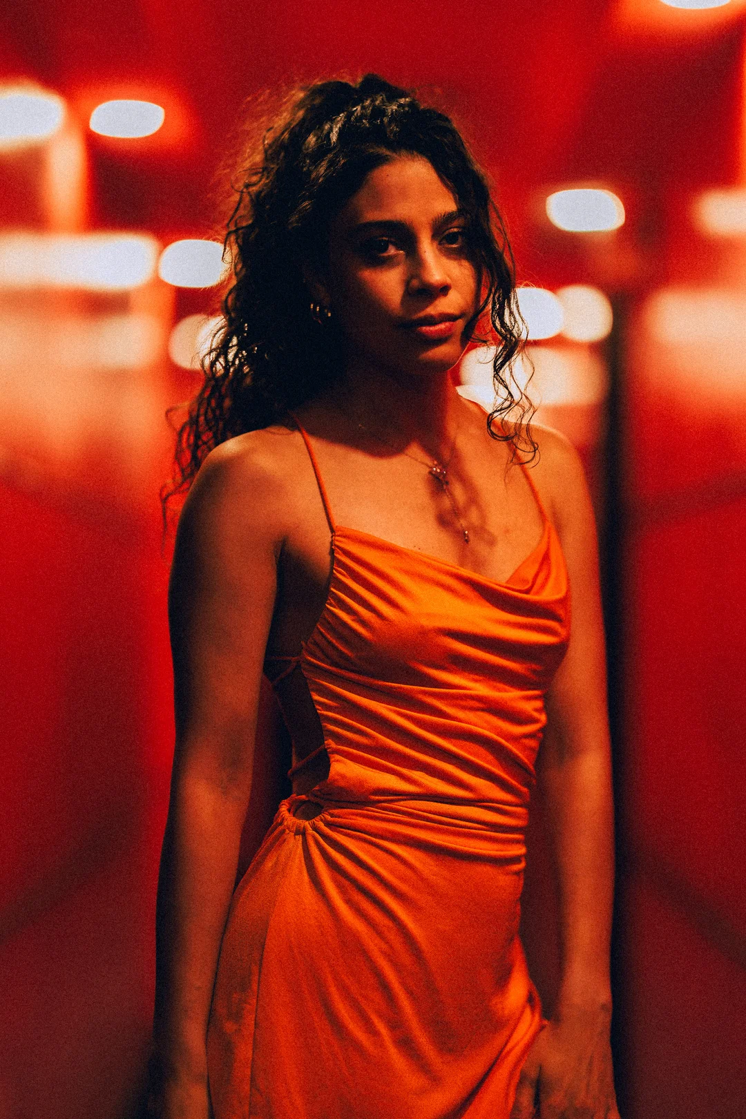Woman with curly hair in an orange satin dress against a moody red background with bokeh lights.