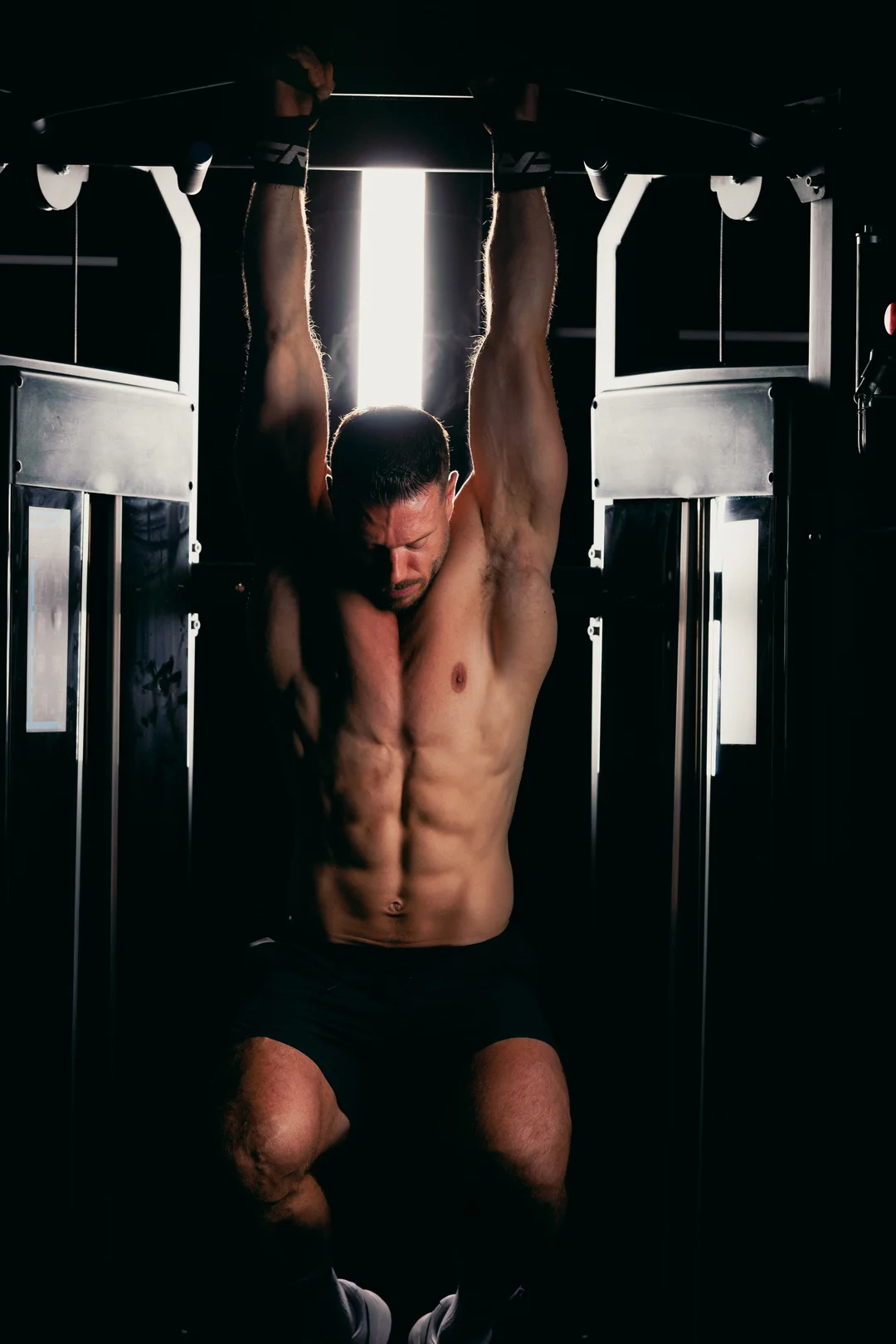 Muscular man performing a pull-up in a gym with dramatic, high-contrast lighting.