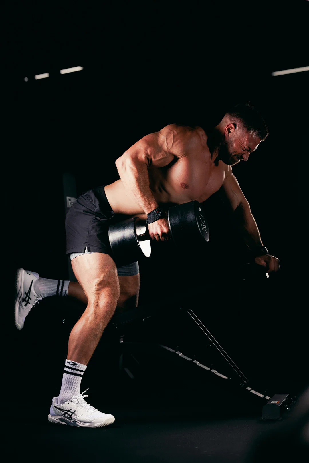Muscular man performing a one-arm dumbbell row in a dark gym with dramatic high-contrast lighting.