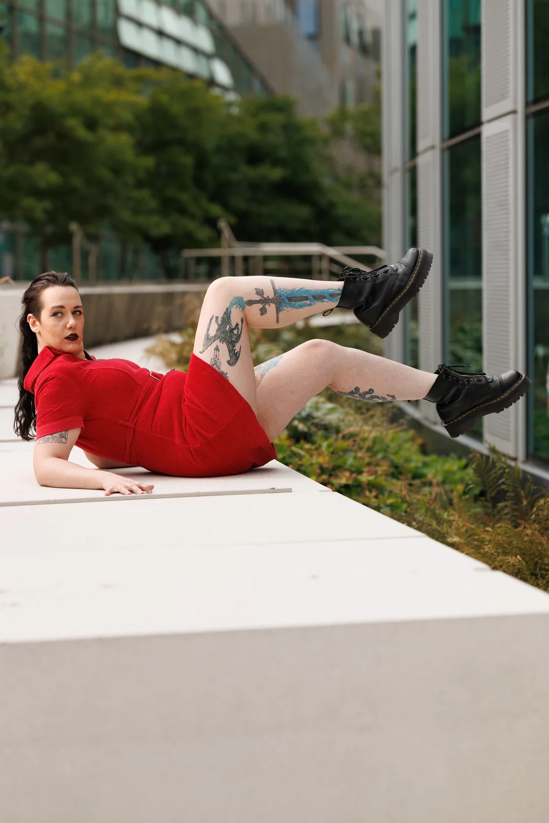 A woman in a red dress and black boots posing on a white concrete ledge in an urban outdoor setting.
